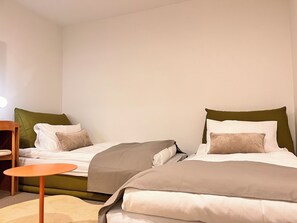 Ein Hotelzimmer mit zwei Betten, jeweils mit einem grünen Kopfteil, weißer Bettwäsche und einem braunen Kissen. In der Mitte des Zimmers steht ein kleiner runder Tisch in Orange.