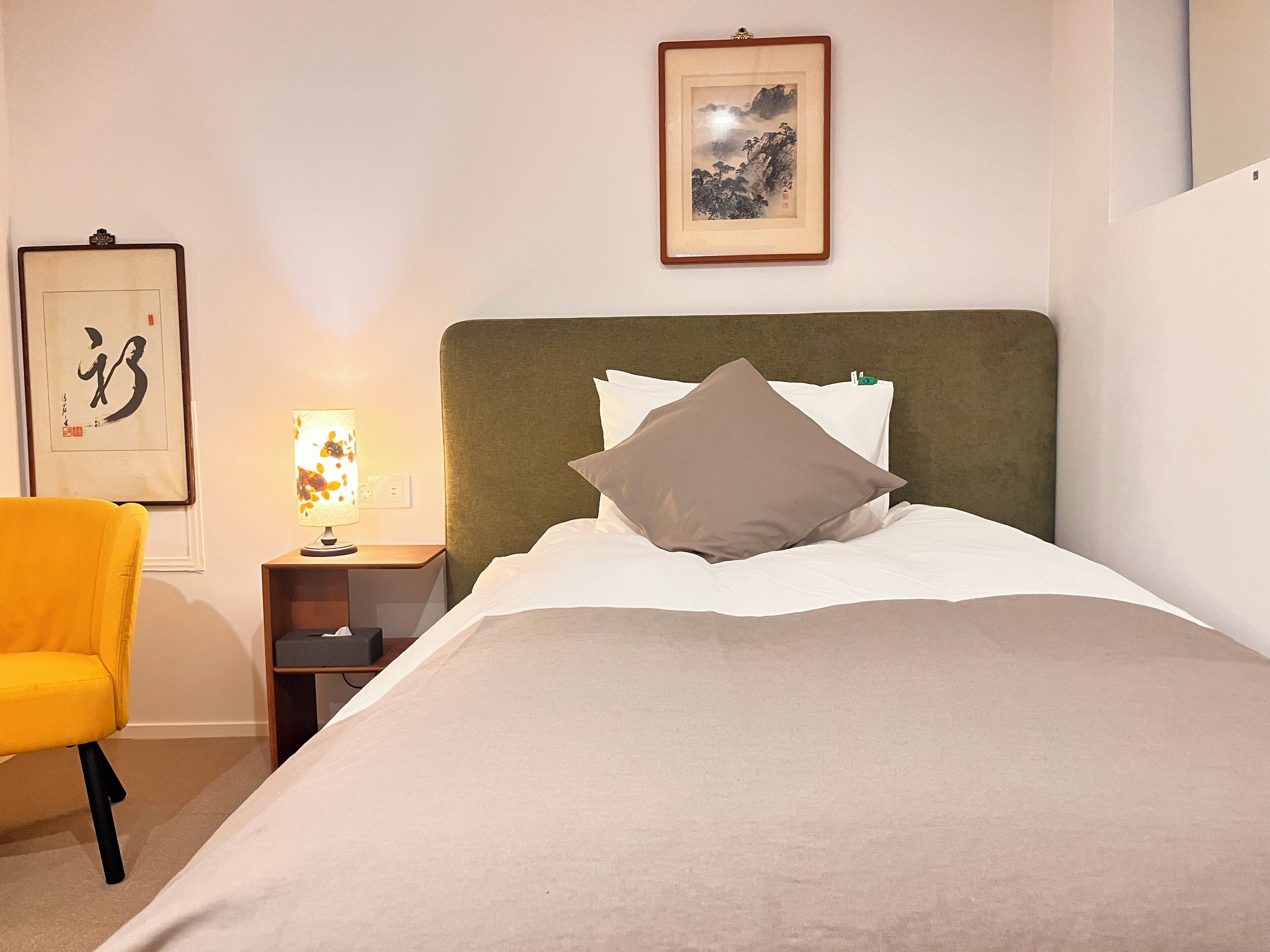 A neatly made bed with a green headboard, a bedside table with a lamp, a framed calligraphy piece, and a yellow chair.