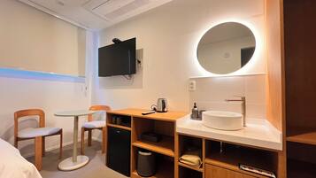 A hotel room with a bed, a desk with a chair, a small table, a TV mounted on the wall, a round mirror above the sink, and a trash bin.