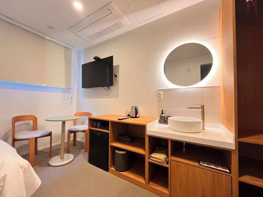 A hotel room with a bed, a desk with a chair, a small table, a TV mounted on the wall, a round mirror above the sink, and a trash bin.