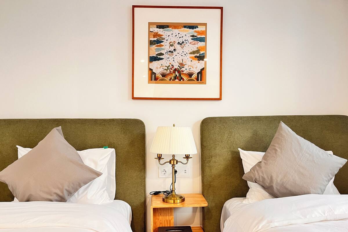 A hotel room with two beds, a nightstand with a lamp, and a framed artwork on the wall.