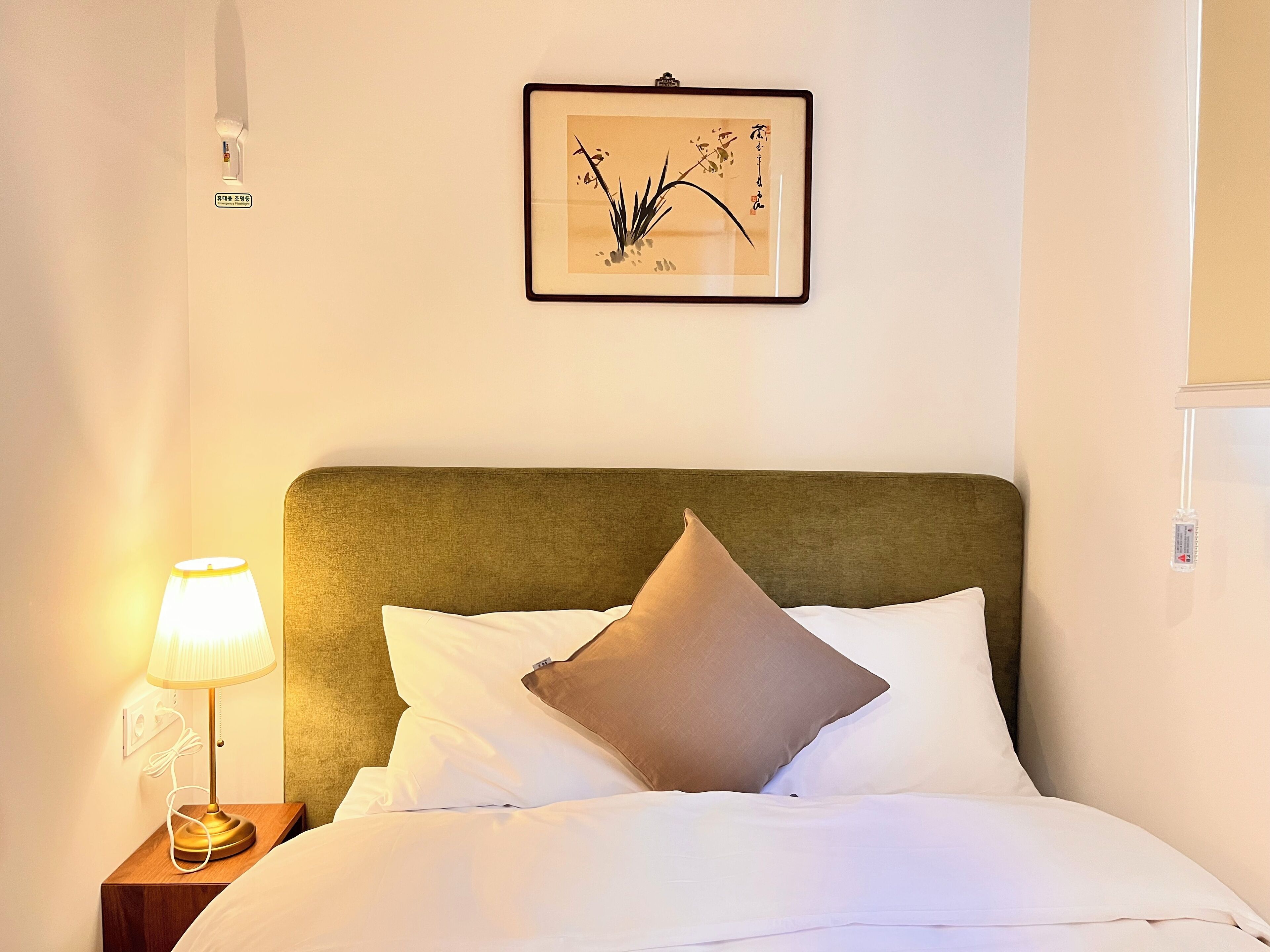 A neatly made bed with white linens and a brown pillow, a bedside lamp, and a framed artwork on the wall.