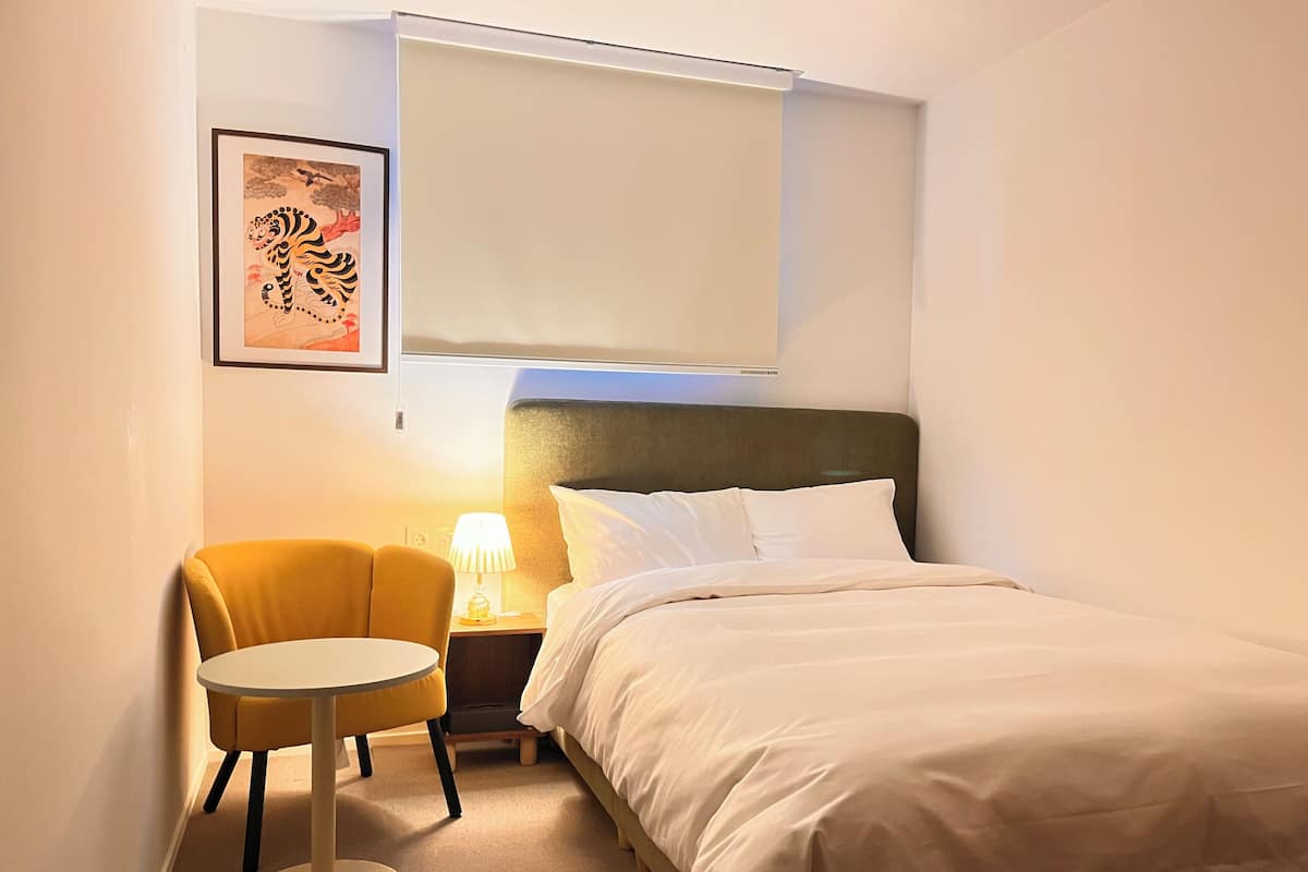 A hotel room with a bed, a chair, a small table, a nightstand, a lamp, and a framed picture on the wall.
