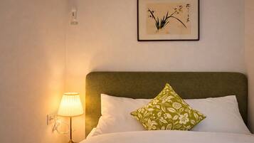 Un letto rifatto con cura, con una testiera verde, un cuscino bianco e un cuscino verde con motivo floreale. A sinistra del letto è presente una lampada fissata al muro.