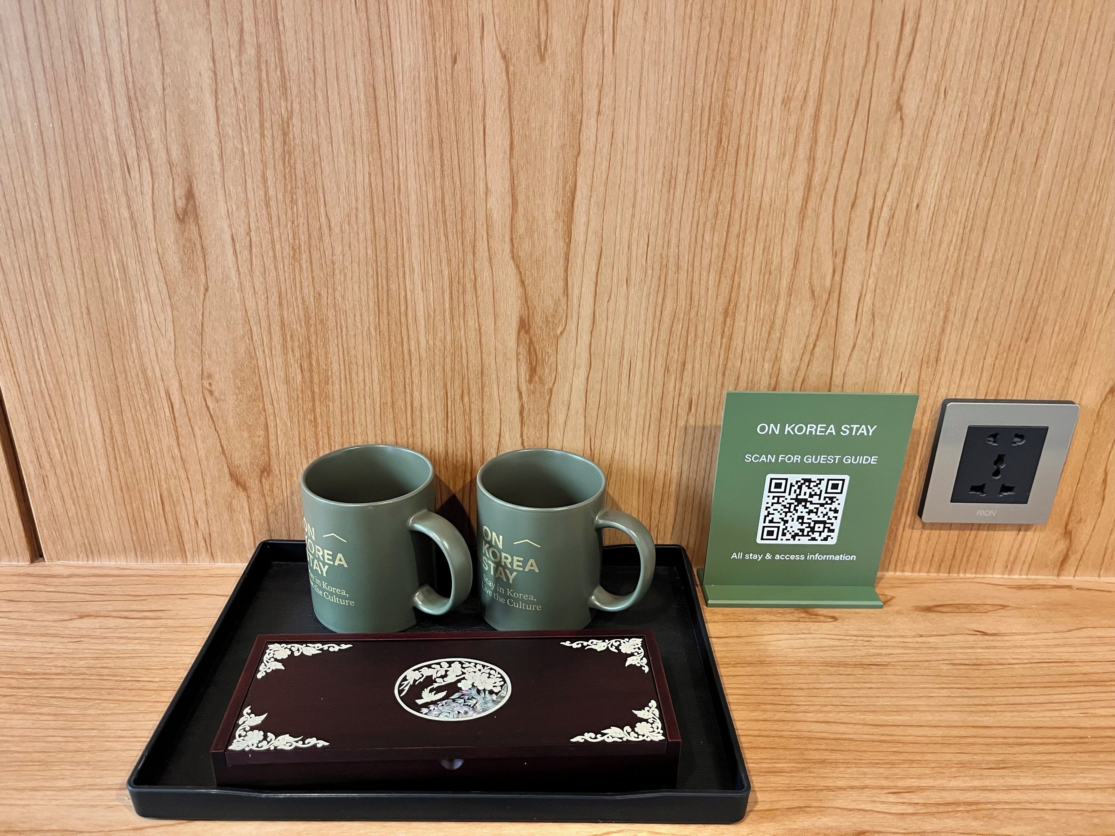 Uma bandeja com duas canecas verdes, uma placa com código QR e uma tomada em uma superfície de madeira.