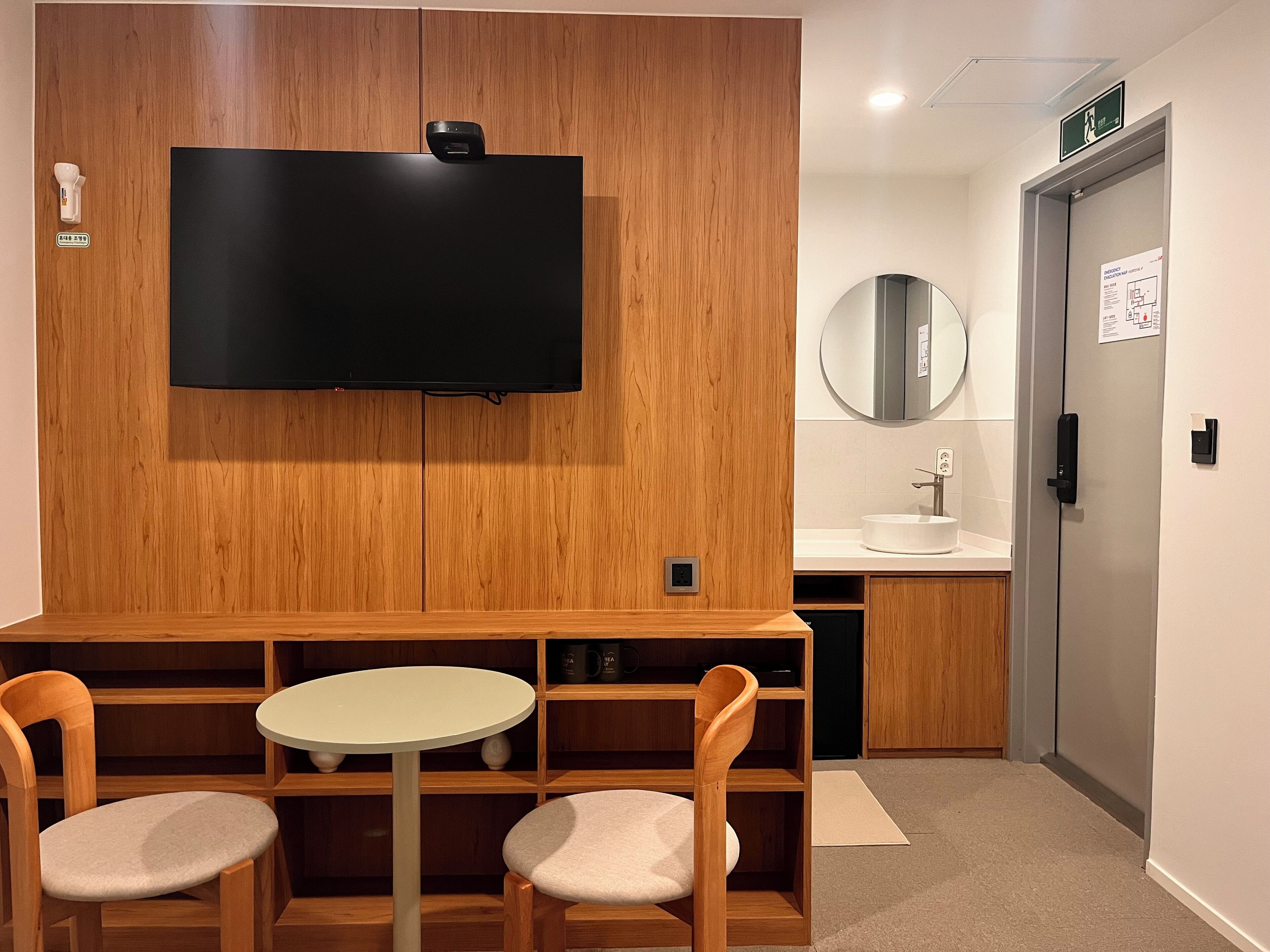 Ein modernes Zimmer mit einem Holzkabinett, einem Flachbildfernseher, einem runden Spiegel, einem Waschbecken und zwei Stühlen um einen kleinen Tisch.