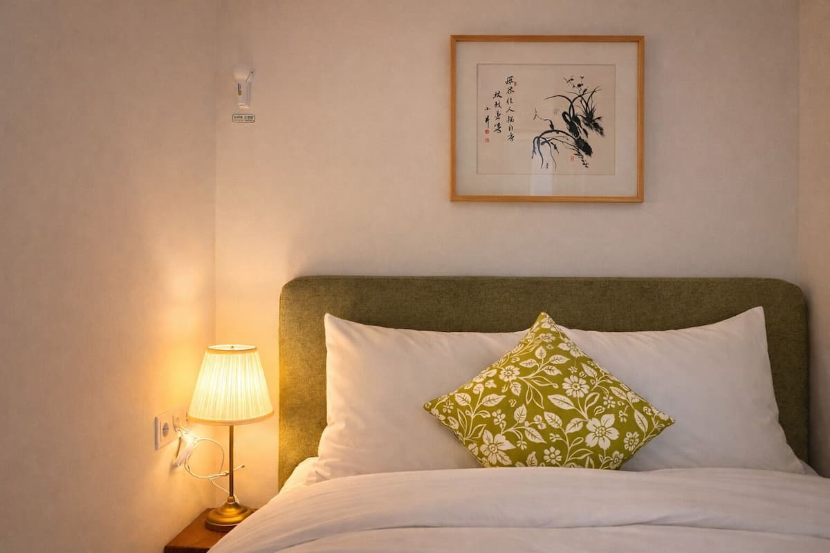 A neatly made bed with white bedding and a green floral pillow, a bedside lamp, and a framed artwork on the wall.