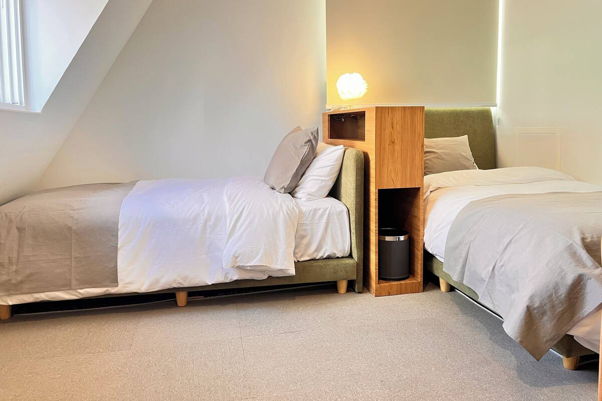 A bedroom with two single beds, a wooden headboard, a bedside table with a lamp, and a window providing natural light.