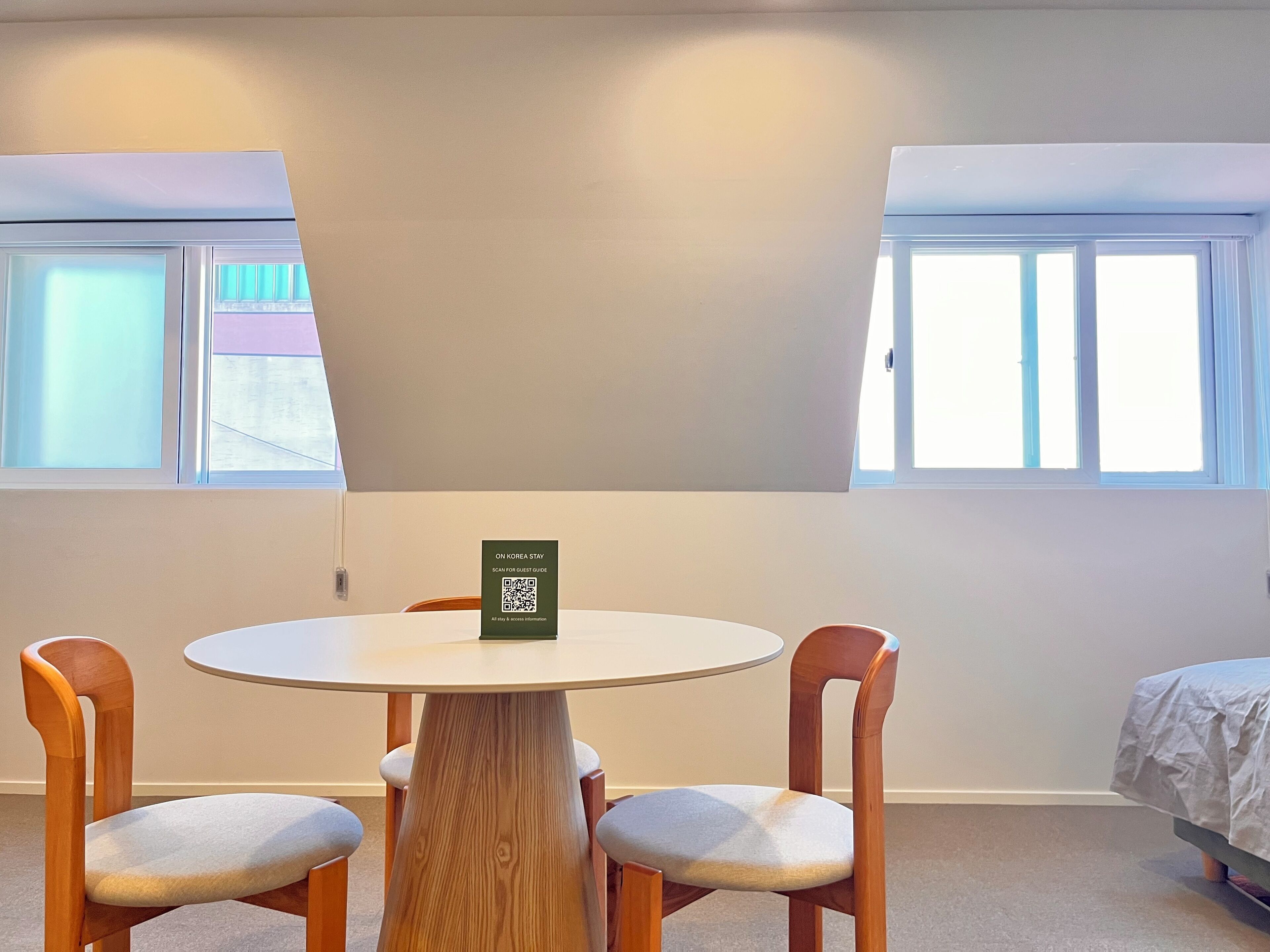 Quarto com uma mesa redonda, duas cadeiras de madeira, uma cama com cobertura branca e uma janela com persianas. Um sinal com código QR está sobre a mesa.