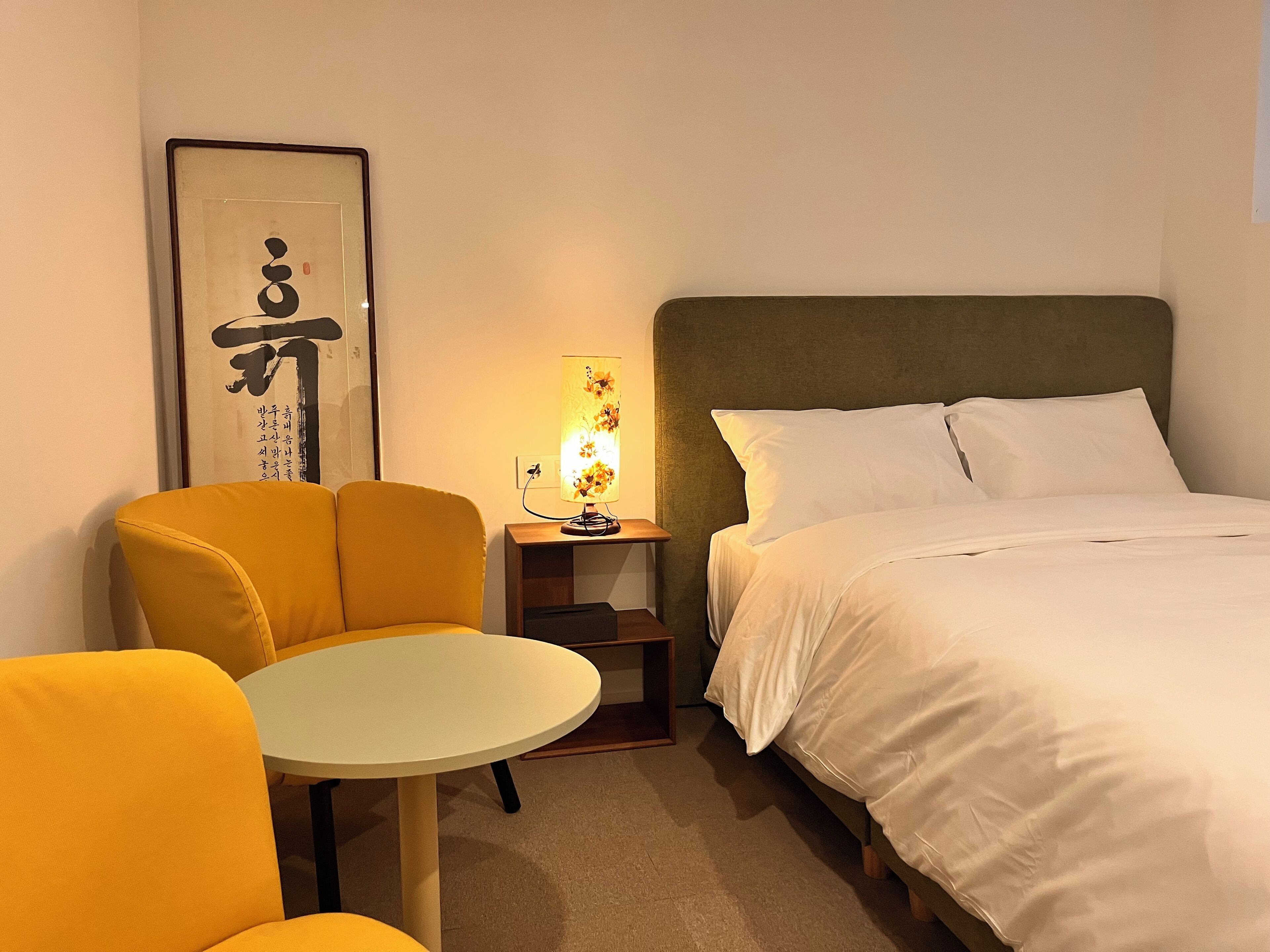 Una habitación de hotel con una cama, una silla, una mesita pequeña y una lámpara fijada a la pared.