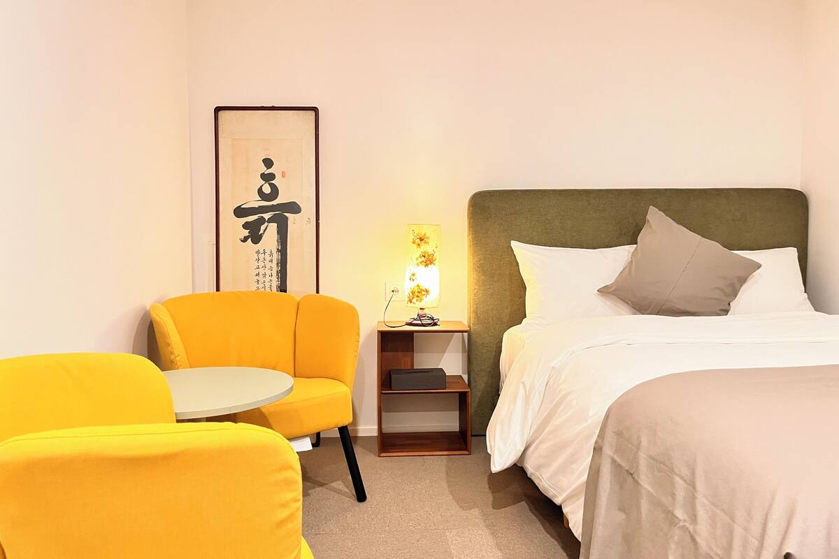 A hotel room with a bed, a yellow armchair, a small table, and a nightstand with a lamp.