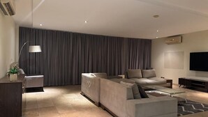 Een moderne woonkamer met een modulaire bank, een glazen salontafel, een televisie en een groot raam met gordijnen.