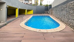 Een rechthoekig zwembad met een houten terras, omringd door een stenen muur en een gebouw met balkons.