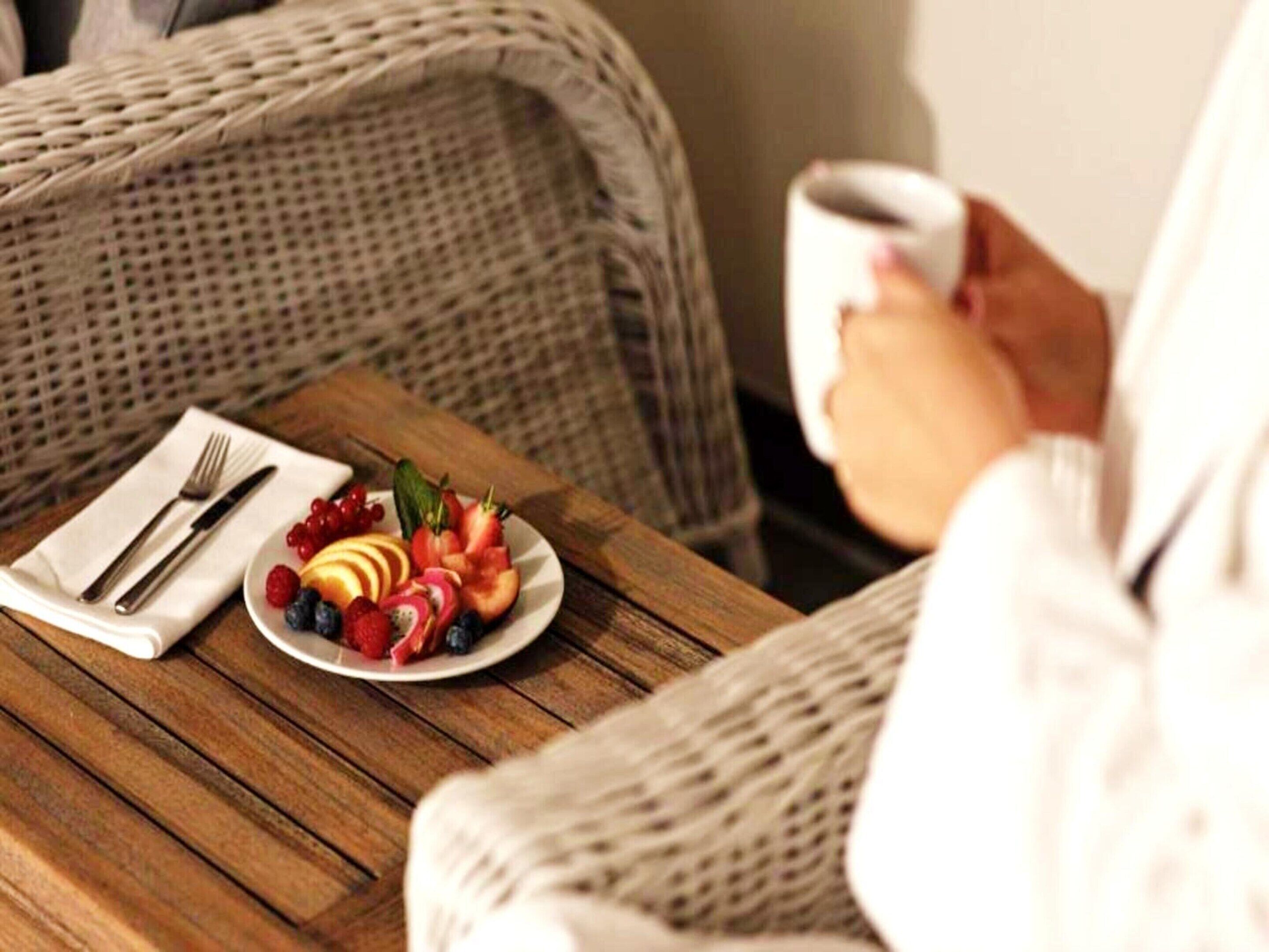 Une chaise en osier, sur une table en bois, avec une assiette blanche remplie de fruits et une tasse de café.