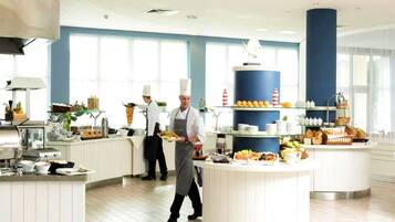 A modern kitchen with chefs preparing food, a buffet area with fruits and pastries, and a large blue pillar.