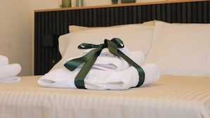 Een net opgemaakt bed met een stapel witte handdoeken samengebonden met een groene lint, een cactussplant en een ingelijst certificaat op het nachtkastje.