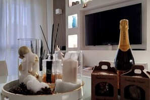 Ein modernes Wohnzimmer mit einem Glastisch, auf dem eine dekorative weiße Skulptur, Kerzen und eine Champagnerflasche stehen. Ein Flachbildfernseher ist an der Wand befestigt, und es befinden sich zwei Kisten Brut Champagner auf dem Tisch.