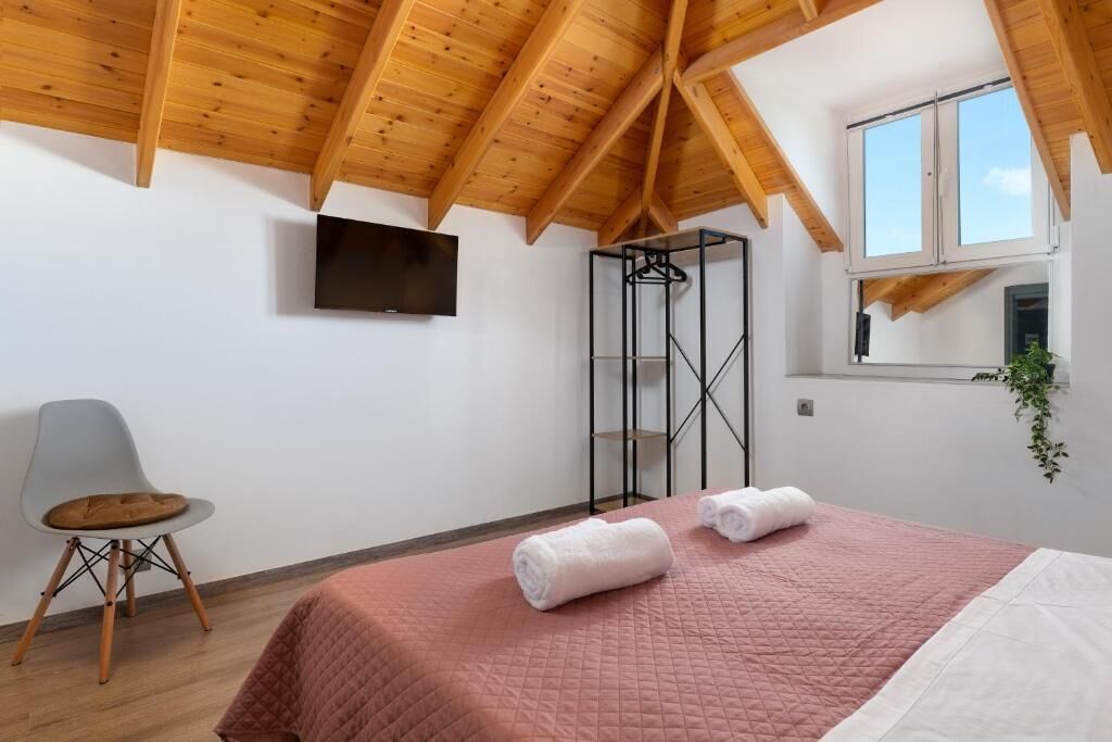 Une chambre avec un plafond en bois, un lit avec une literie rose, un téléviseur fixé au mur et une fenêtre avec une plante.