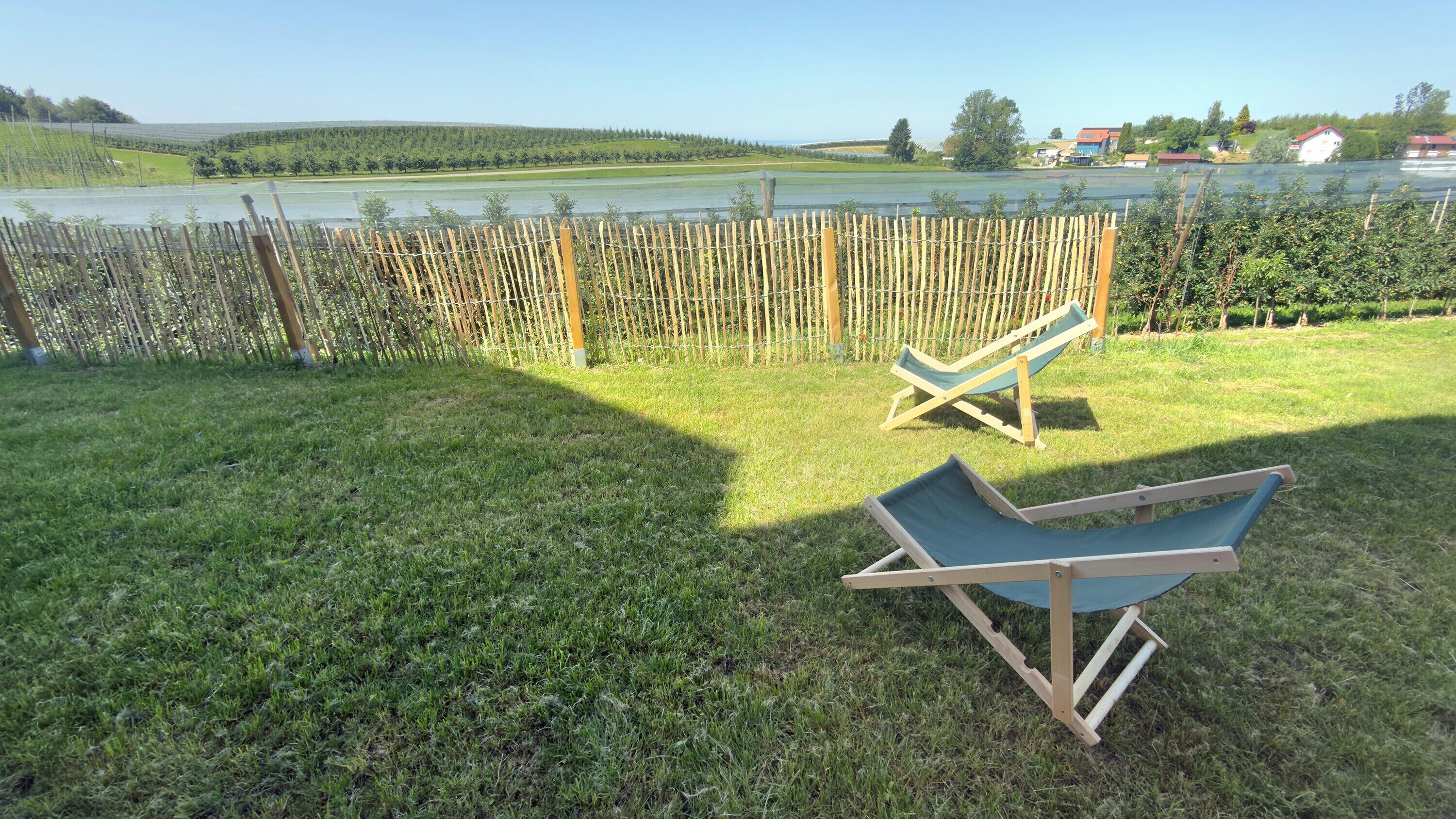 Ein grüner Hinterhof mit Holzzaun, zwei Holzliegen und Blick auf einen See sowie entfernte Gebäude.