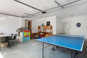 Ein Raum mit Tischtennisplatte, Stühlen, einem kleinen Spielhaus, Bücherregalen und einer Dartscheibe.