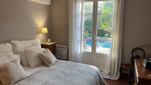 Ein Schlafzimmer mit Bett, Nachttisch, Lampe und Blick auf einen Pool durch ein Fenster.