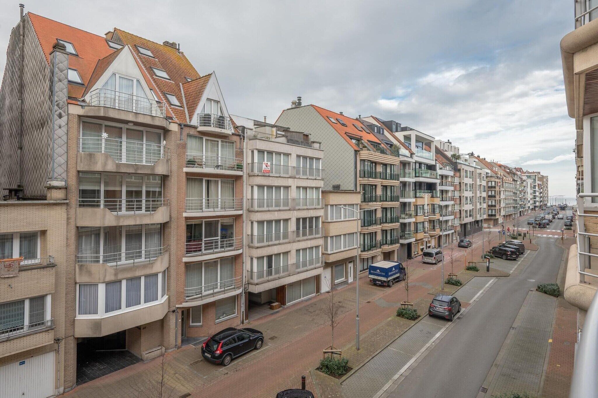Een rij moderne appartementencomplexen met balkons, een verharde straat en geparkeerde auto's.