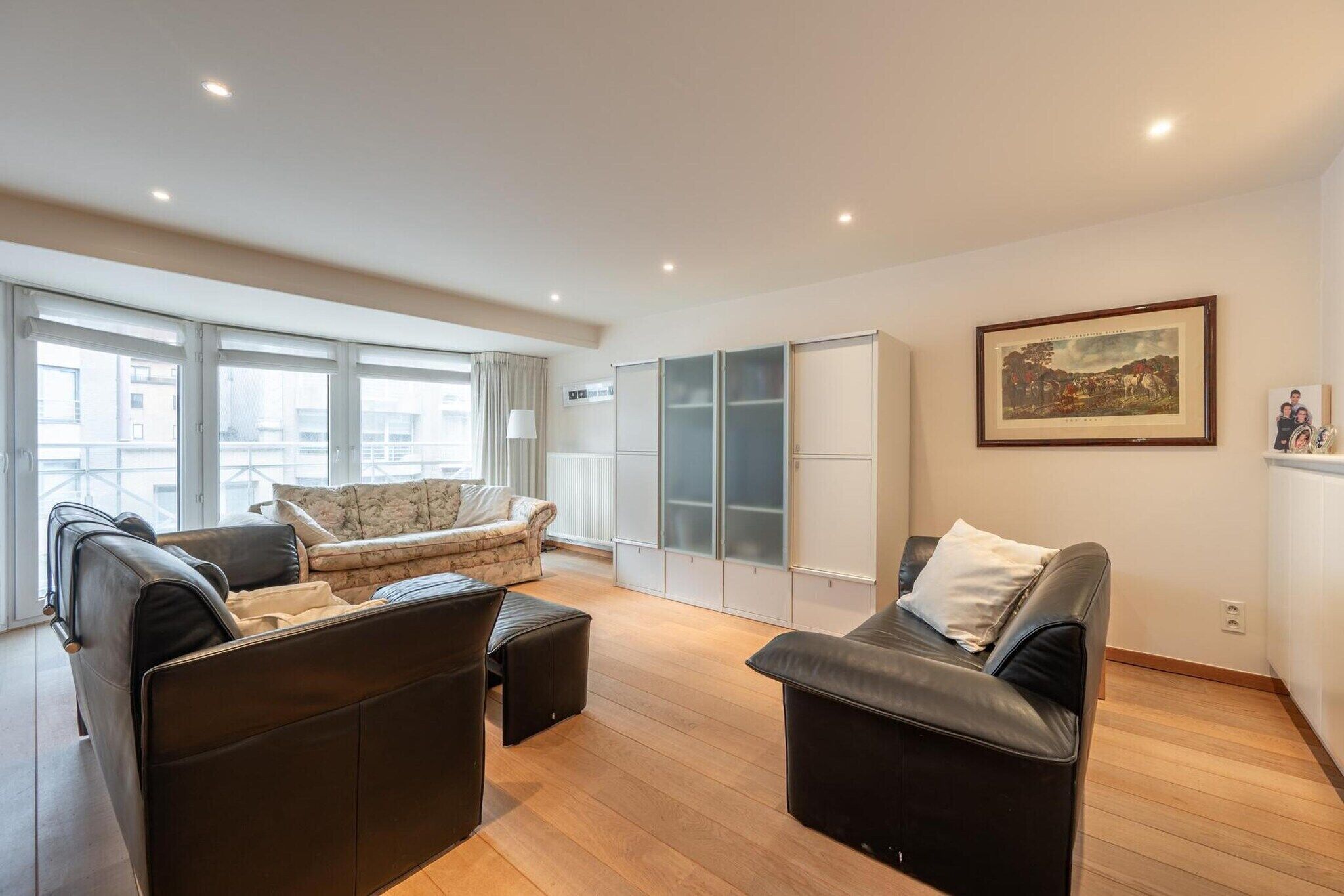 A spacious living room with a sofa set, a coffee table, and a dining area with a table and chairs. There is a large window providing natural light and a view of the outside.