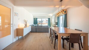 A modern living room with a sofa set, a wooden dining table, and a large window with curtains. - 3-Bedroom Sunny Apartment in Knokke (KNOKKE-HEIST)