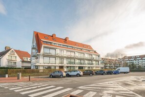 En moderne, flereetagers boligbygning med rødt tag, flere balkoner og en parkeringsplads med biler og en varevogn.