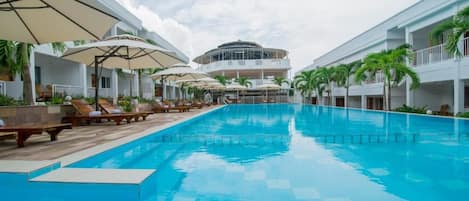 Una piscina con sillones de descanso y sombrillas, rodeada de edificios con balcones y palmeras.