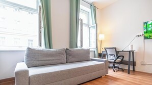 Een moderne woonkamer met een grijze bank, een bureau met stoel, en een aan de muur bevestigde televisie.