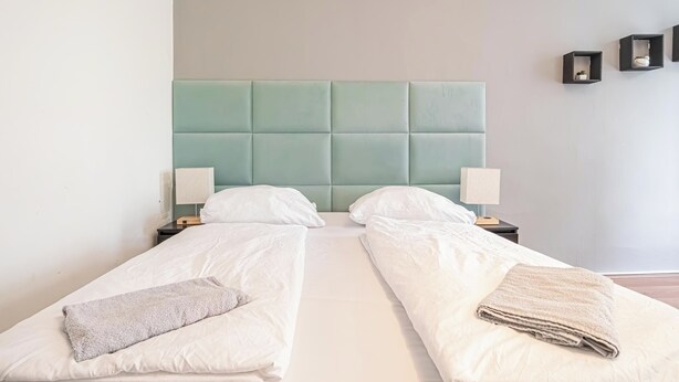 Een tweepersoonsbed met een lichtgroen hoofdeinde, twee nachtkastjes met lampen, en een wandplank met decoratieve objecten.