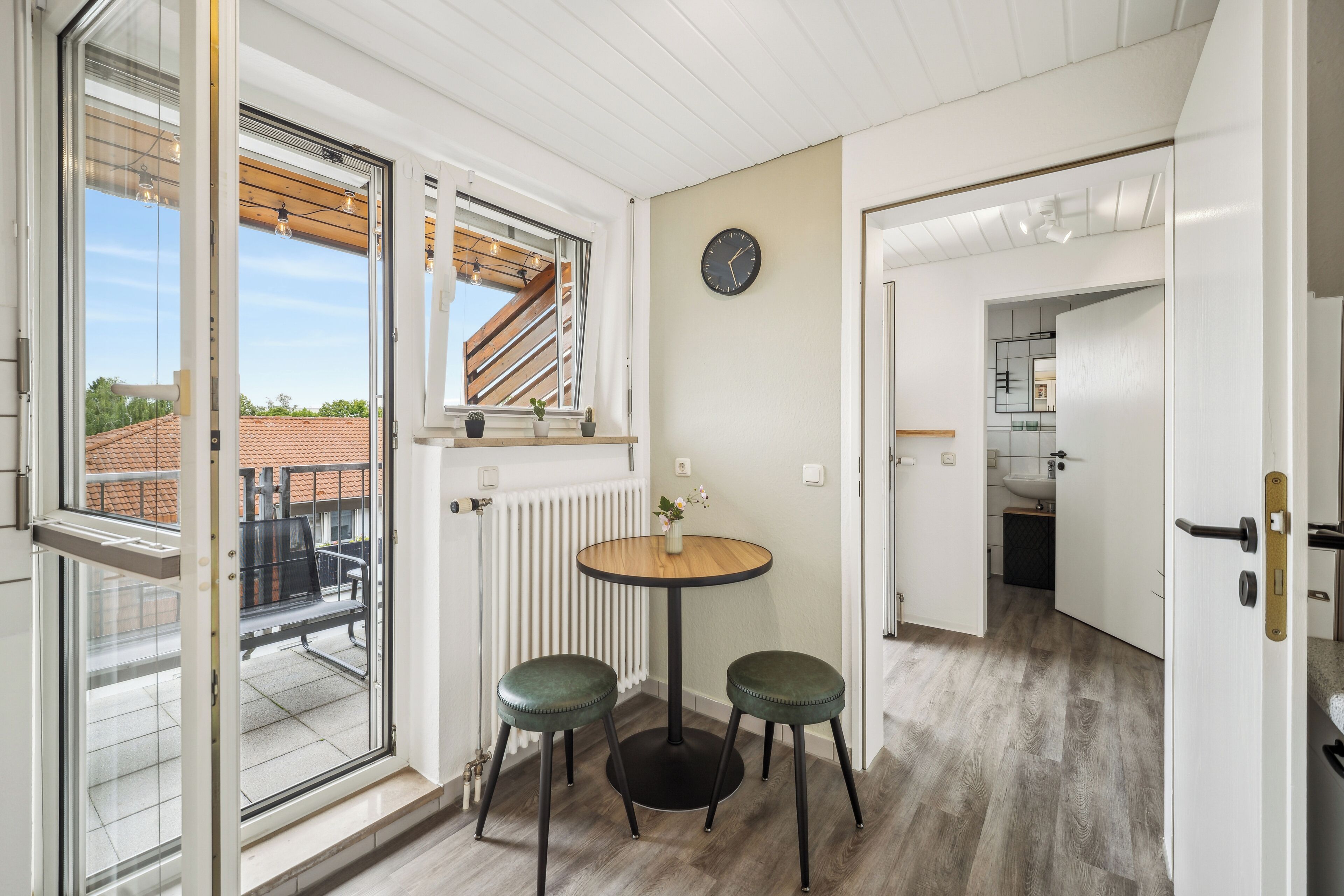 Ein Zimmer mit einer Glastür zum Balkon, einem runden Holztisch mit zwei grünen Hocker, einer Uhr an der Wand und einer Heizung.