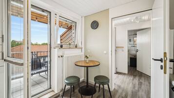 Ein Zimmer mit einer Glastür zum Balkon, einem runden Holztisch mit zwei grünen Hocker, einer Uhr an der Wand und einer Heizung.