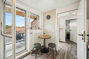 Ein Zimmer mit einer Glastür zum Balkon, einem runden Holztisch mit zwei grünen Hocker, einer Uhr an der Wand und einer Heizung.