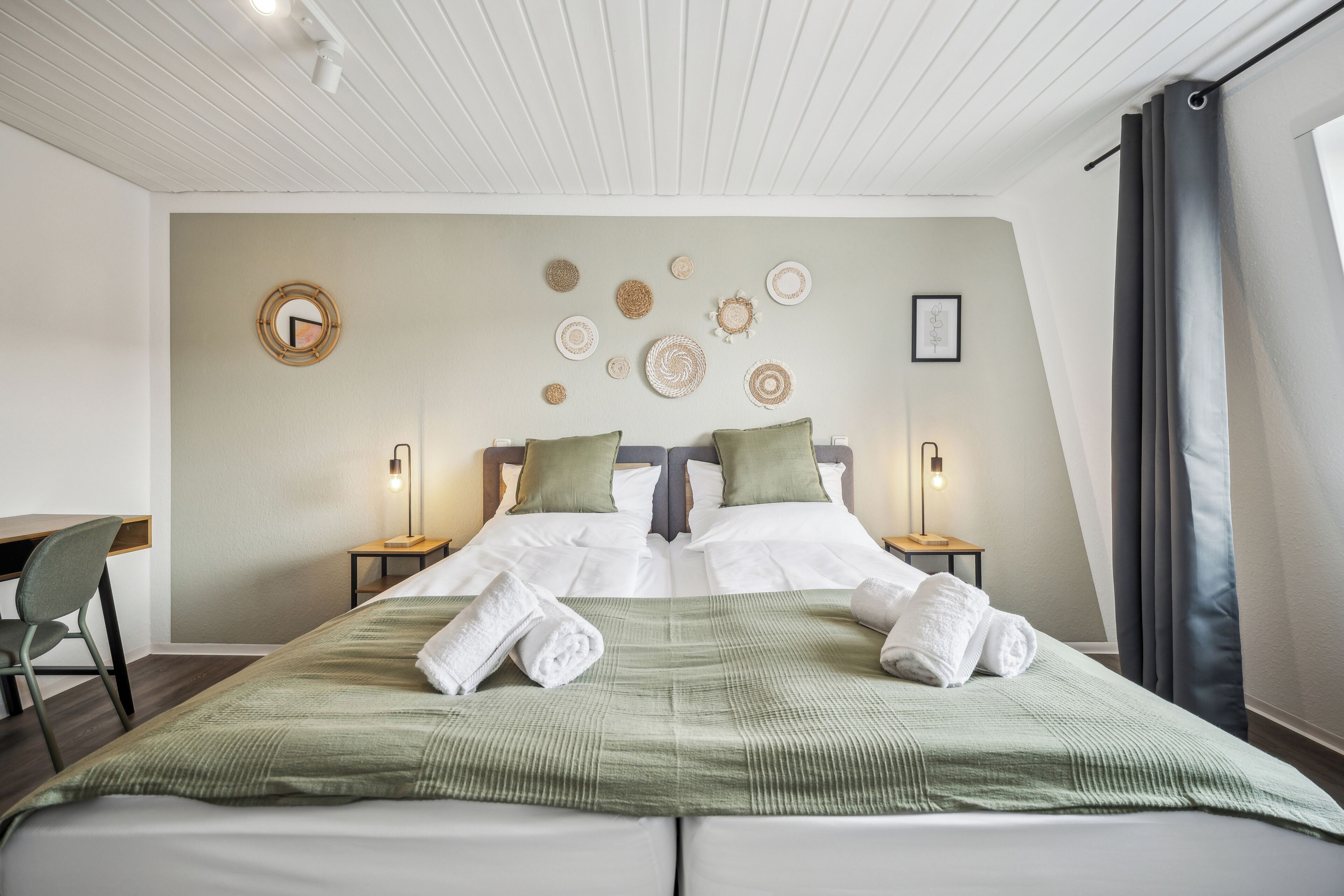 Ein ordentlich bezogenes Doppelbett mit grüner Bettwäsche, zwei Nachttische mit Lampen, ein Schreibtisch und ein Stuhl in einem modernen Zimmer mit schrägen Wänden und dekorativer Wandkunst.