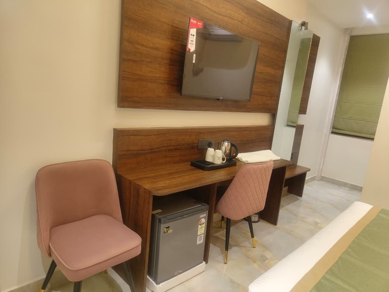 A compact hotel room with a wooden desk, two pink chairs, a small refrigerator, a flat-screen TV mounted on the wall, and a green window.
