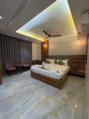 Een moderne slaapkamer met een groot bed, een plafondventilator, een houten hoofdbord en een zithoek met stoelen.