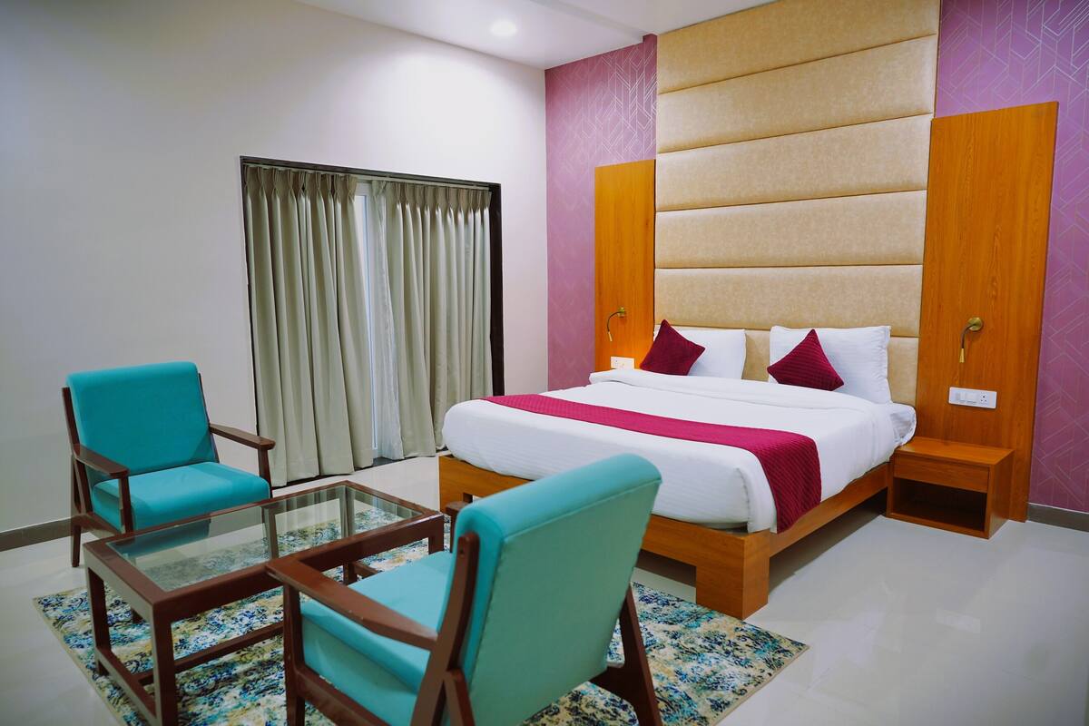 Una habitación de hotel con cama, dos sillas, una mesa de vidrio y una ventana con cortinas.