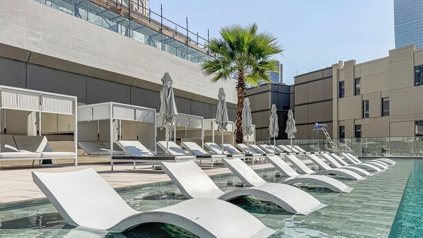 Zona de piscina con tumbonas y sombrillas, una palmera y un edificio moderno al fondo.