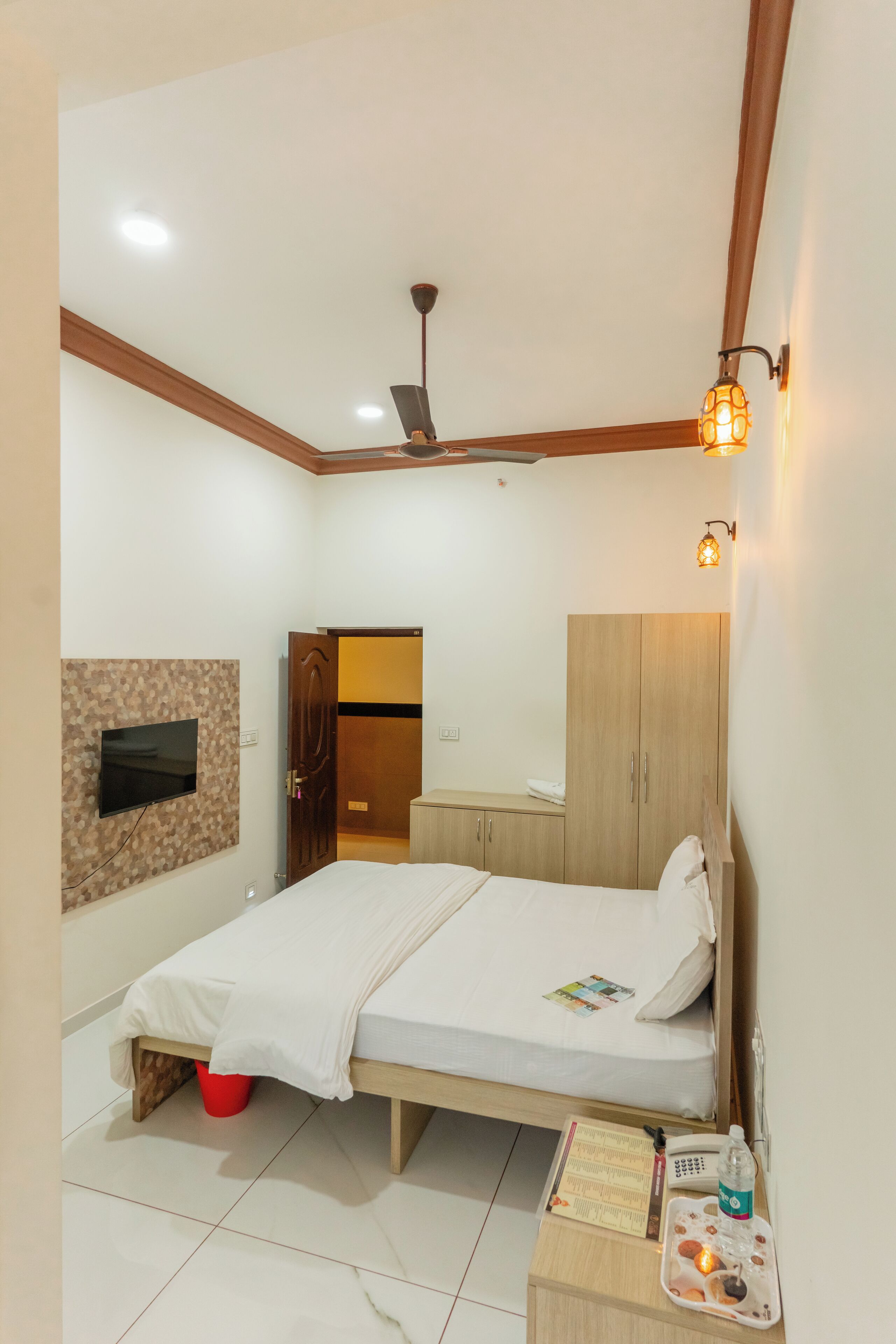 A hotel room with a bed, a TV mounted on the wall, a wardrobe, a bedside table with a phone and a bottle, and a ceiling fan.