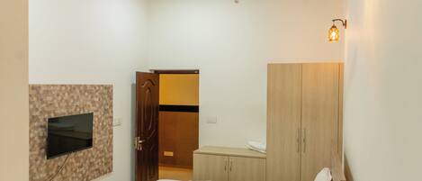 A hotel room with a bed, a TV mounted on the wall, a wardrobe, a bedside table with a phone and a bottle, and a ceiling fan.