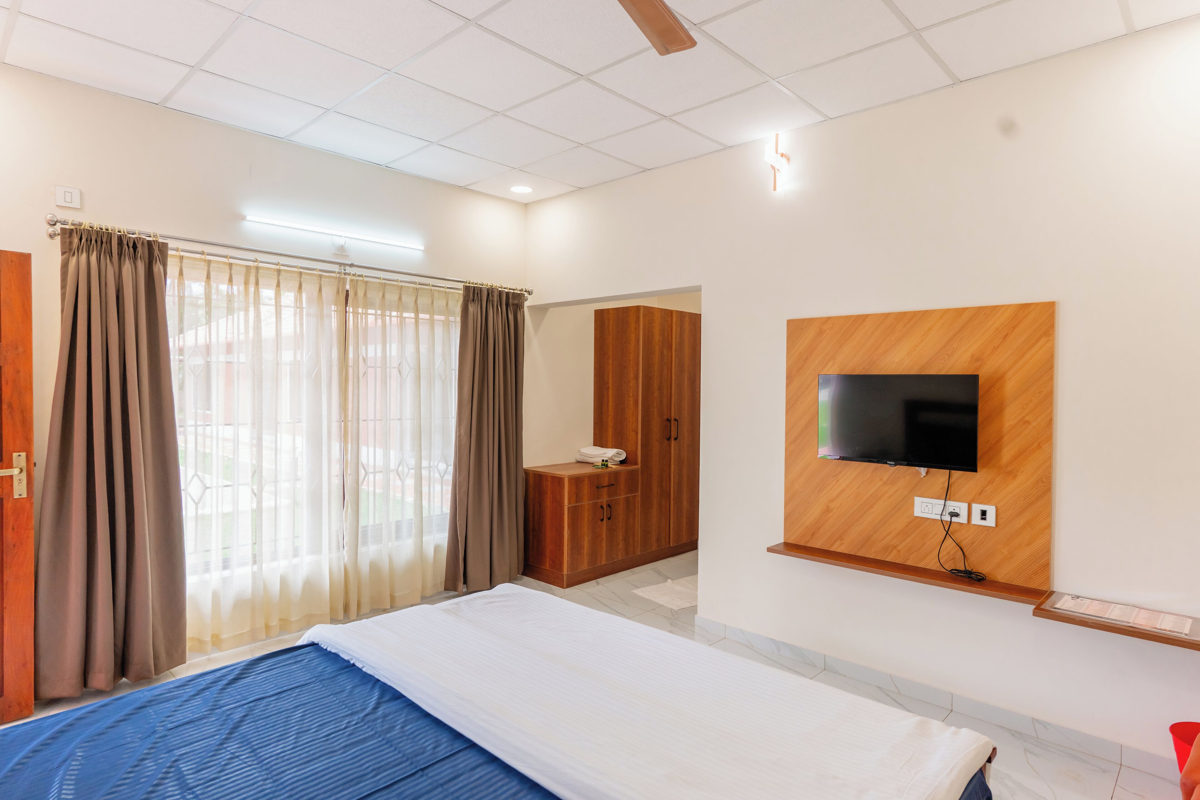A hotel room with a bed, a TV mounted on the wall, a wardrobe, and a window with curtains.
