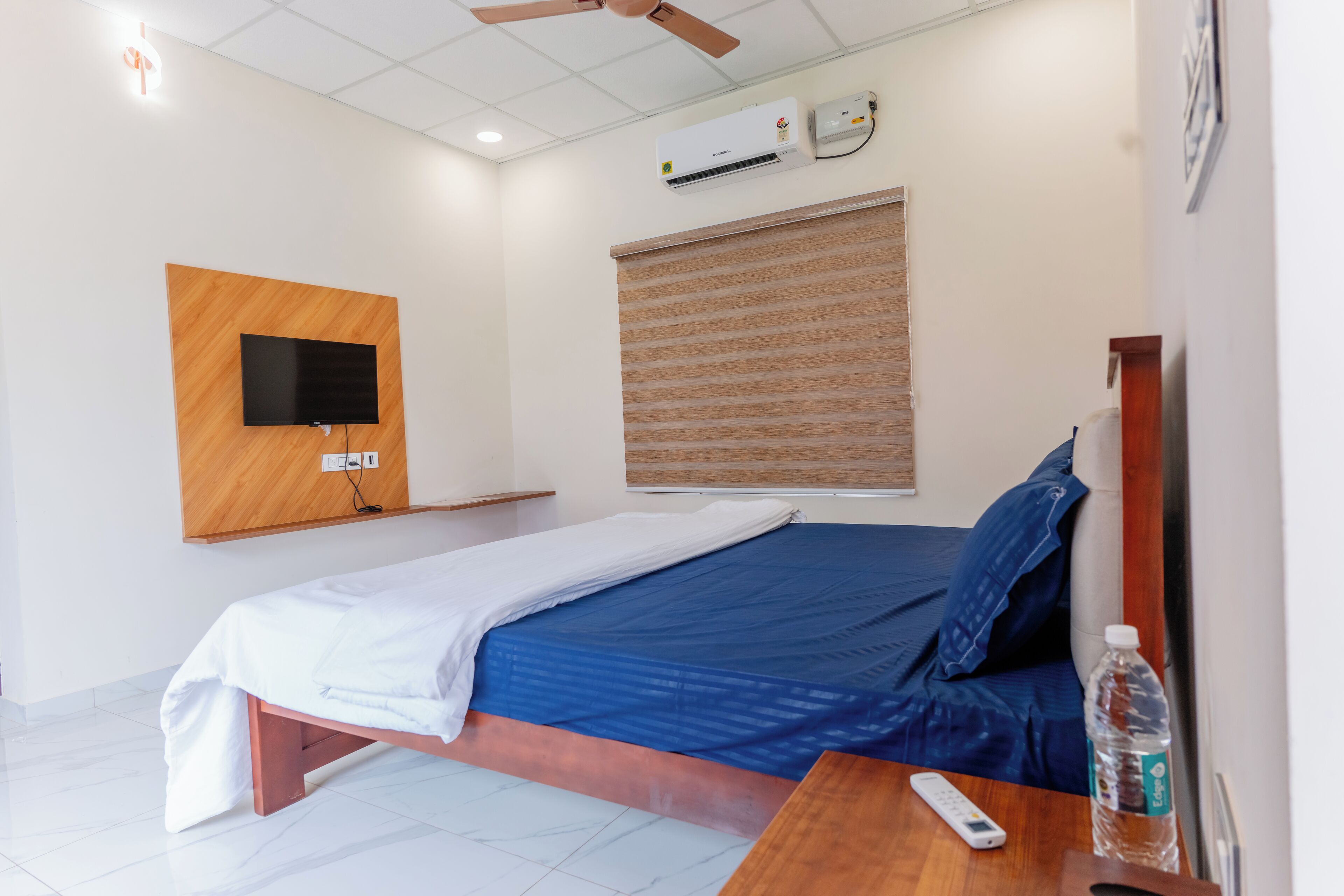 A single bed with a blue mattress, a wooden headboard, a mounted TV, a ceiling fan, and an air conditioning unit.