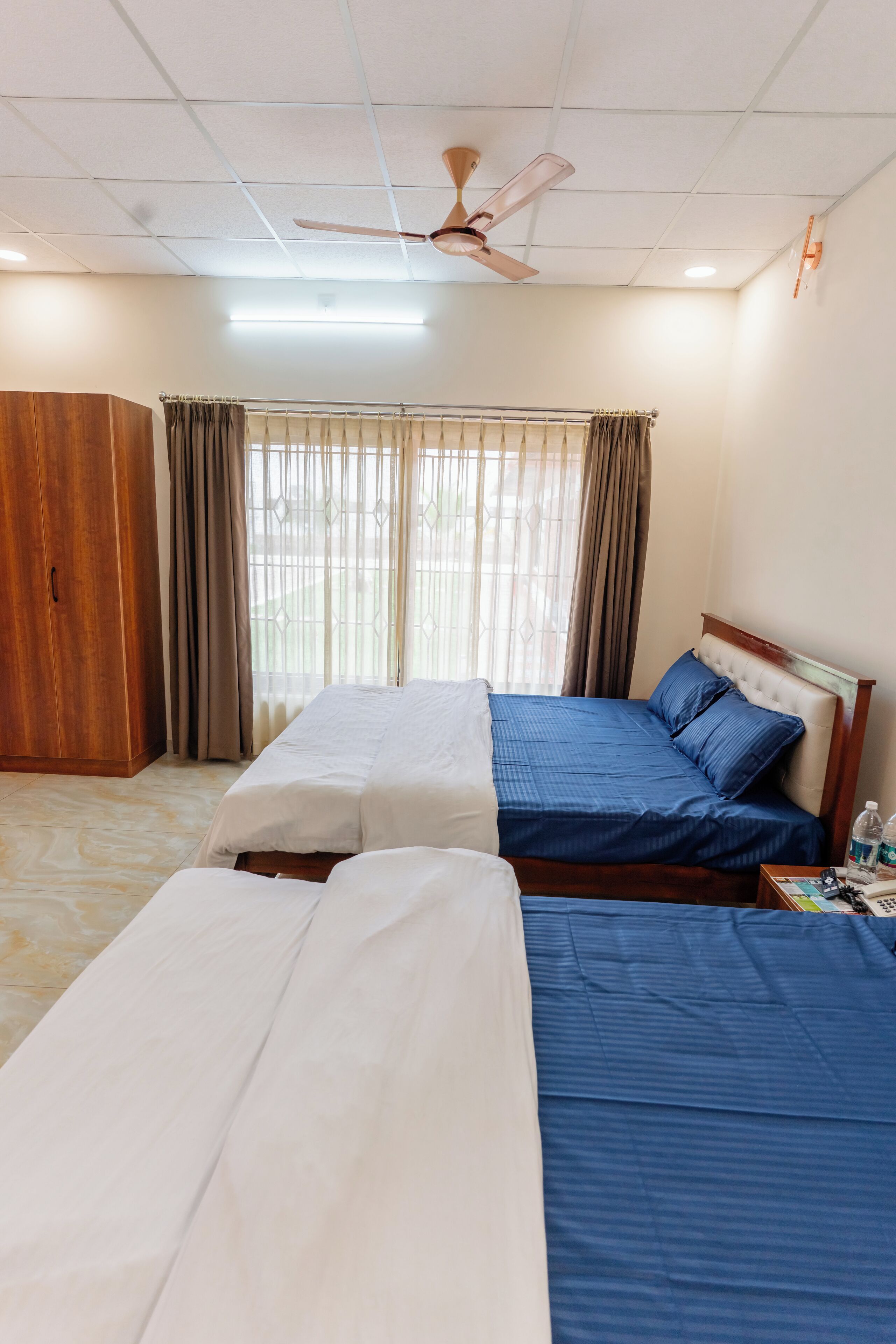 A hotel room with two beds, a wardrobe, a ceiling fan, and a window with curtains.
