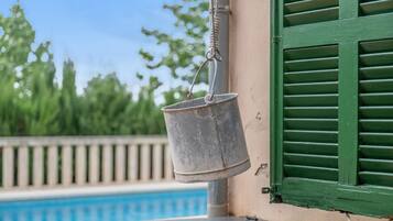A metal bucket hanging from a pole by a rope, a pool, and a building with green shutters.