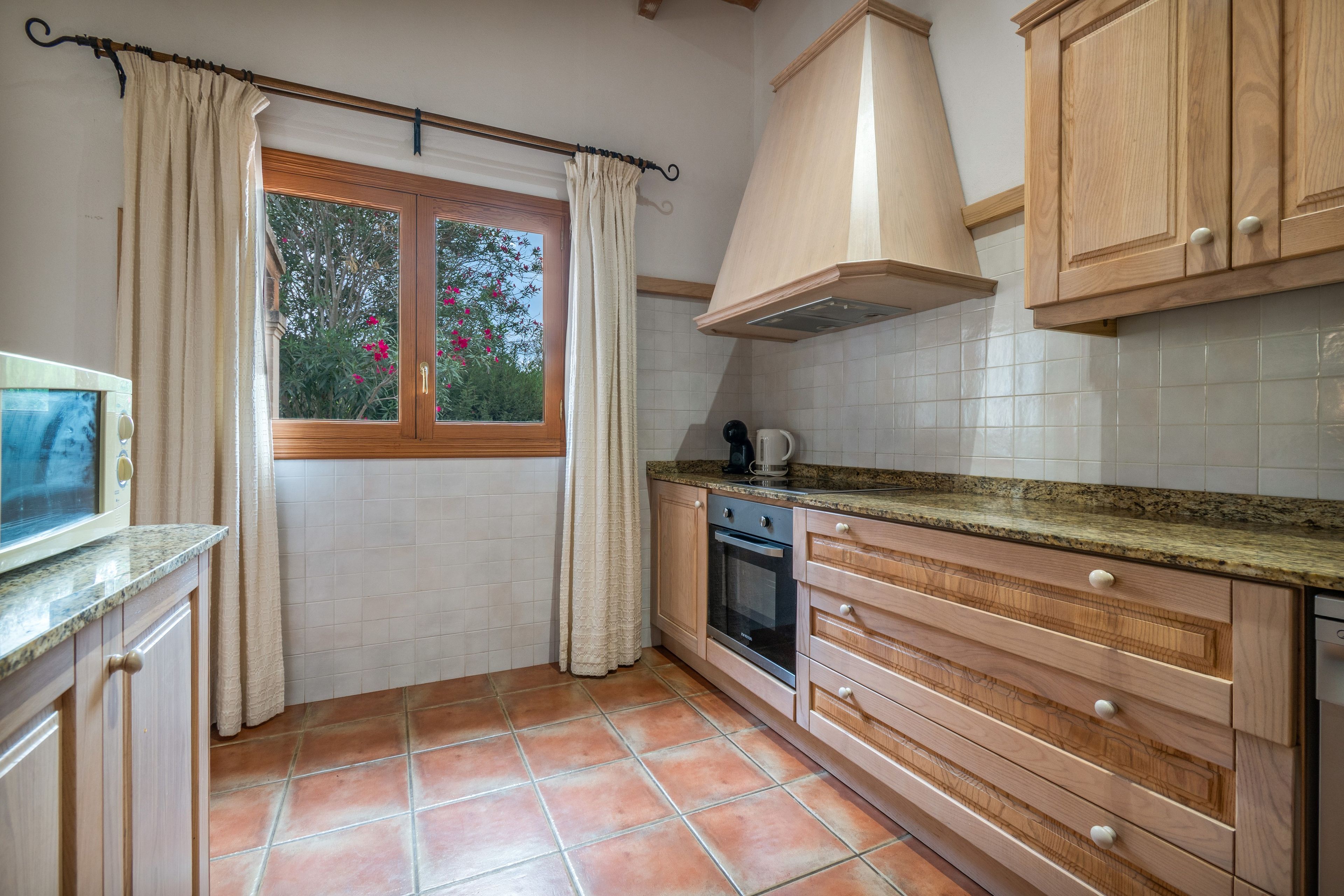 Eine Küche mit Holzausstattung, einer gefliesten Spritzschutzwand, einem Fenster mit Vorhängen und einem gefliesten Boden.