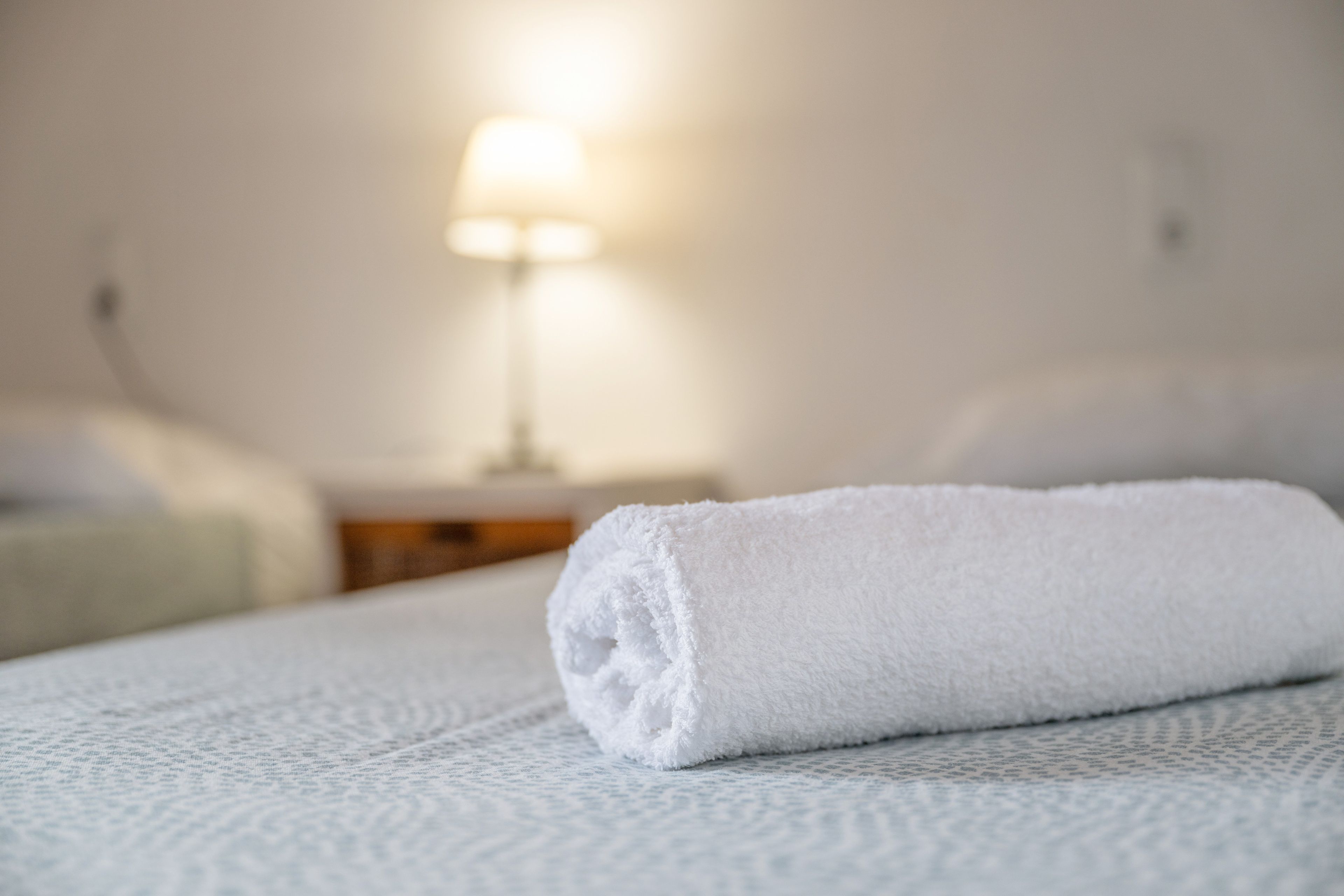 Une serviette blanche, soigneusement pliée, est posée sur un lit recouvert d’une couverture blanche à motifs.