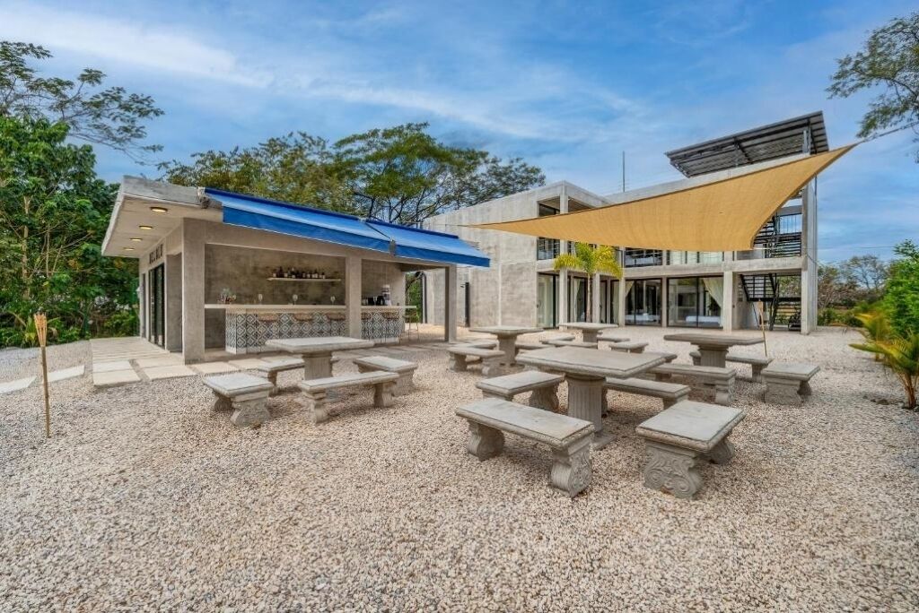 An outdoor patio area with stone tables and benches, a covered seating area, and a building with a blue roof.