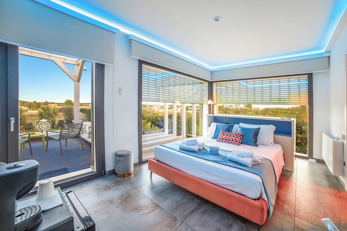 A modern bedroom with a large bed, a view of the outdoors, and a balcony with outdoor furniture.